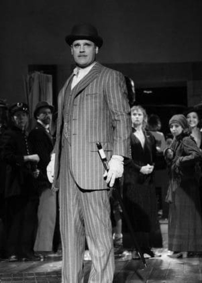 Man in pinstripe suit from "The Threepenny Opera" with a group of actors in the background