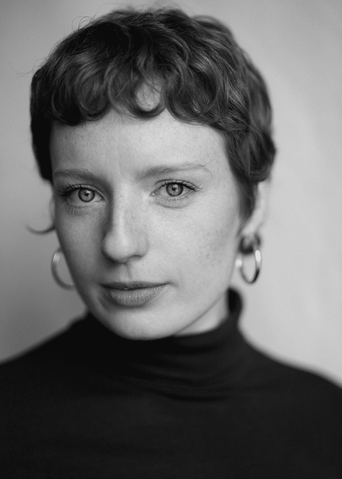 Portrait of Tilly Botsford with short, curly hair, wearing a black turtleneck and hoop earrings.