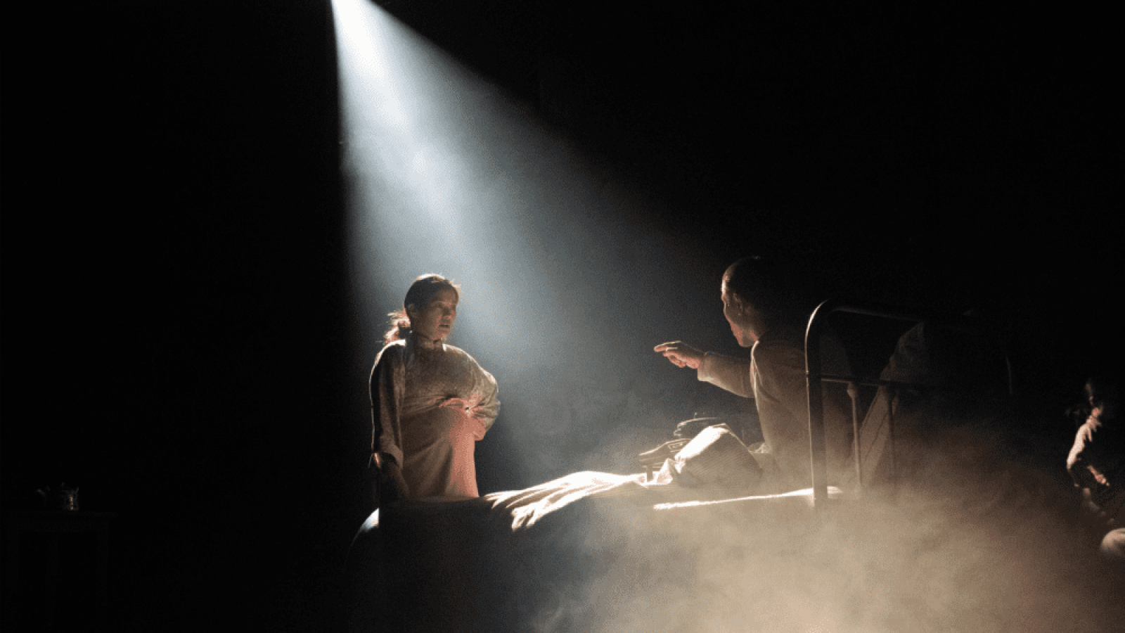 A woman and man converse intensely in a dimly lit room, a single spotlight illuminating them. The far country setting is suggested by the mood.
