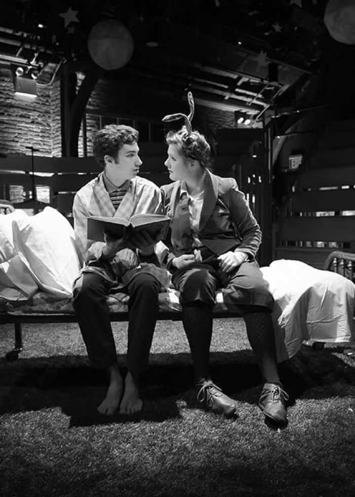 Two actors in The Velveteen Rabbit production sit on a bed, reading a book, with musicians in the background.