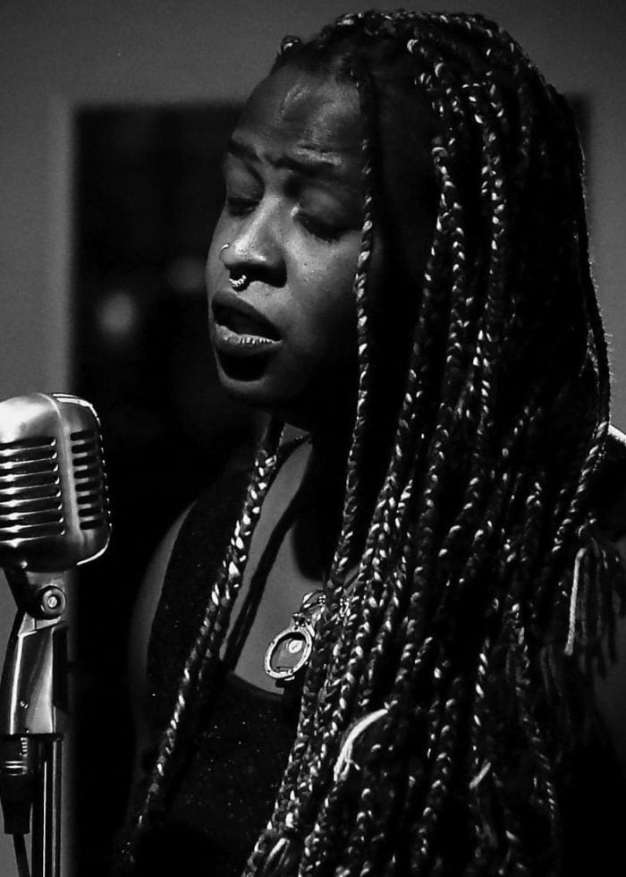 Vita E. Cleveland singing into a vintage microphone, wearing braids and a pendant.