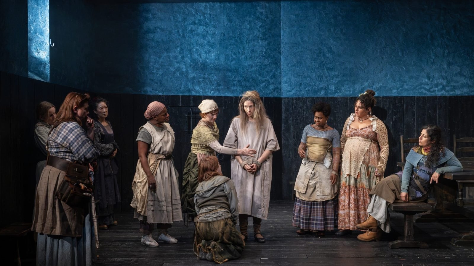 A group of women in period costumes gather on a dark stage, one kneeling in the center.