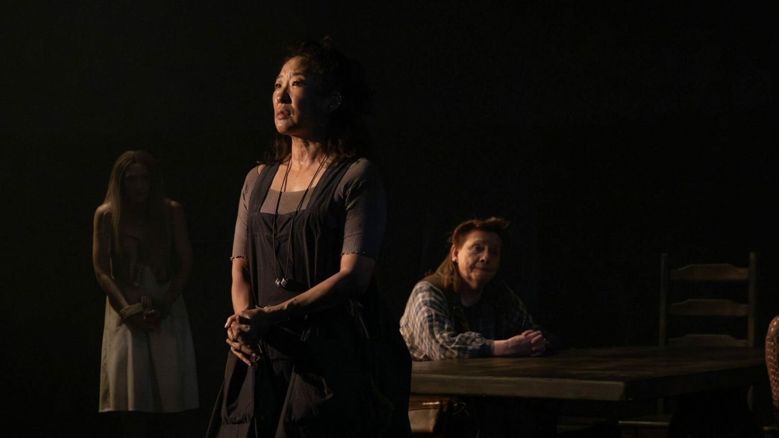 A woman stands center stage, gazing upward, as other women sit at a table in a dimly lit room. The Welkin.
