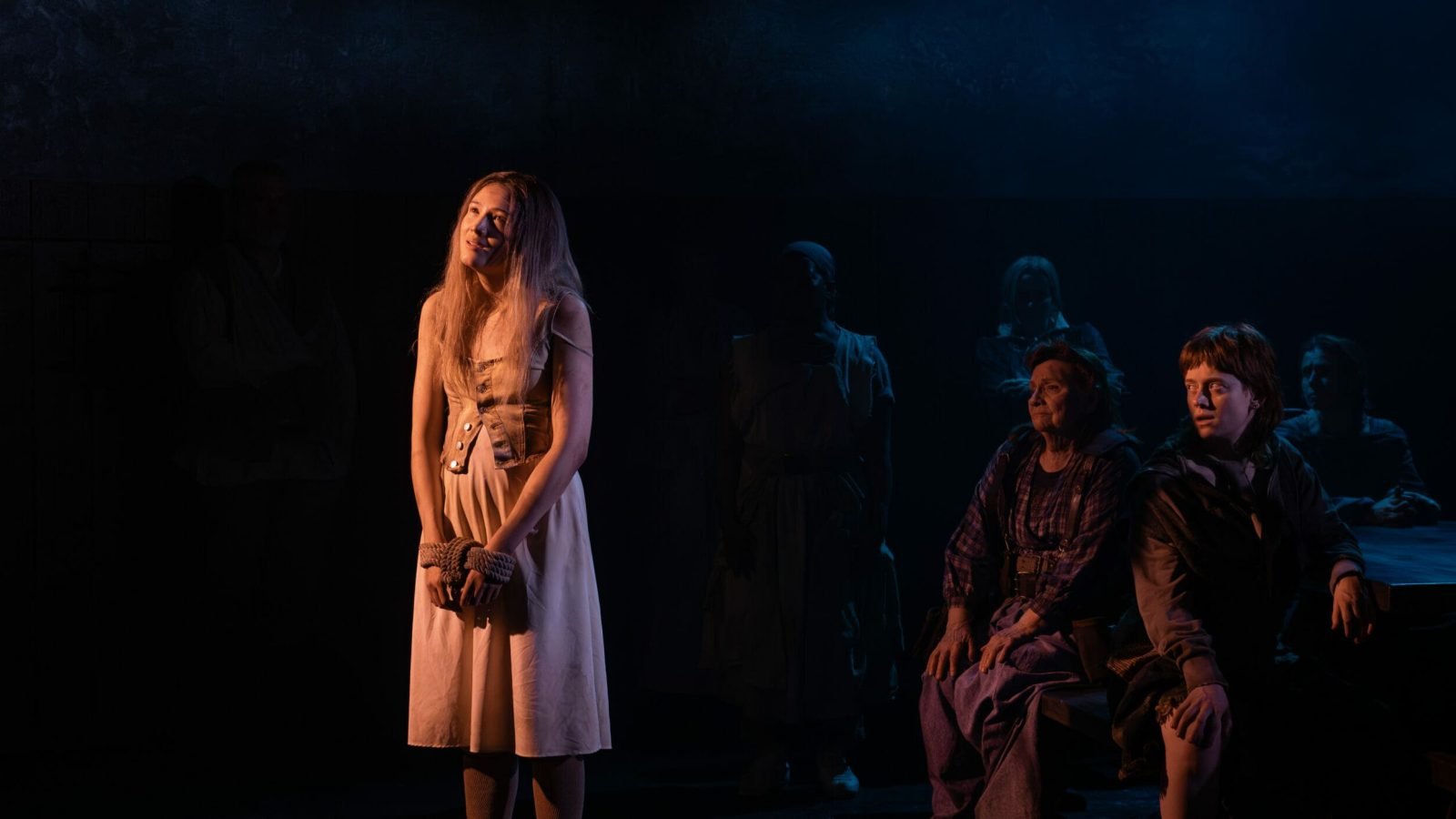 A young woman, hands bound, stands center stage in a dimly lit theatrical setting, looking upward. Other figures are seated in the background.