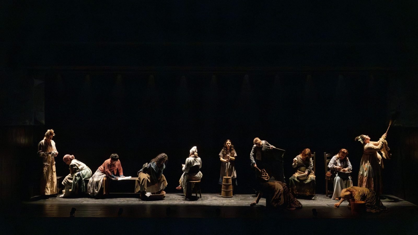 A group of women in period costumes perform a scene on a dark stage.