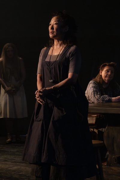 A woman in a dark dress stands center stage, hands clasped, gazing upward. Two other figures are visible in the background.