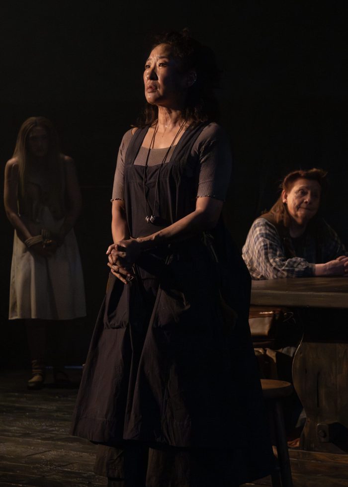 A woman in a dark dress stands center stage, hands clasped, gazing upward. Two other figures are visible in the background.