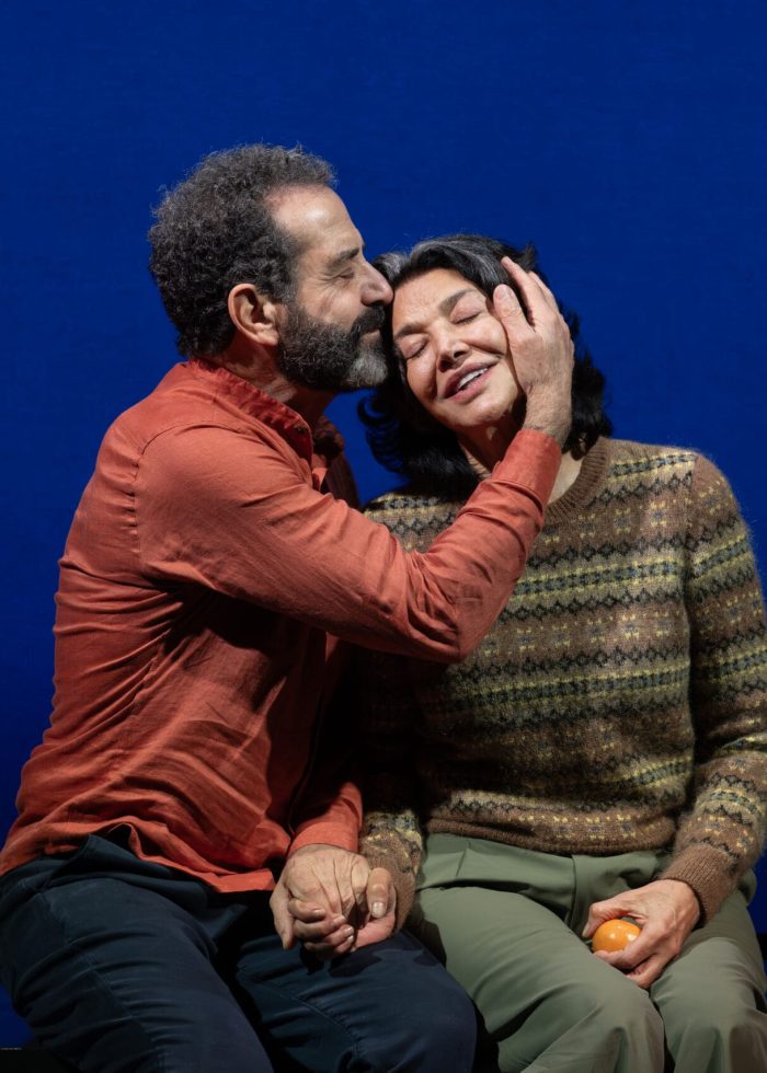 Man kisses woman's head, holding hands, with an orange. Intimate moment on a blue backdrop.