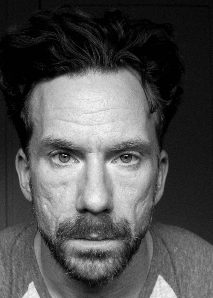 Close-up black and white portrait of a man with a beard, looking directly at the camera.