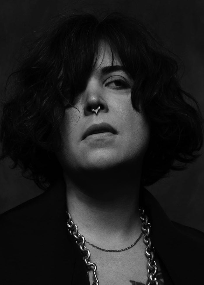 Black and white portrait of a person with a nose ring and chain necklace, looking directly at the viewer.