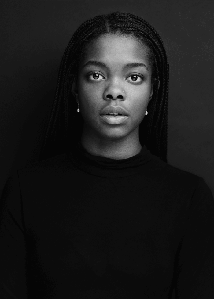Portrait of a young Black woman with long braided hair, wearing a black turtleneck.