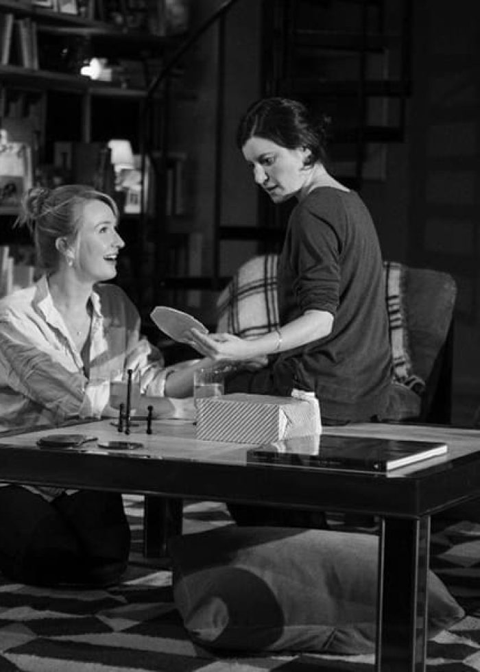Two women in a theater production, seated near a table, discussing a prop. Review of Women or Nothing.
