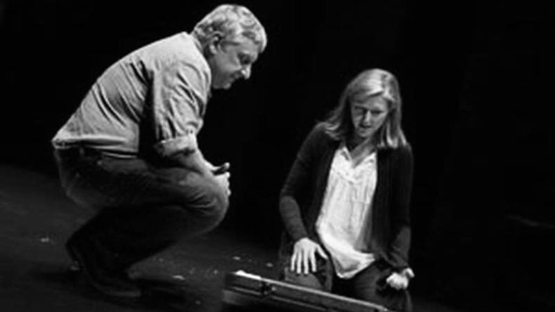 Actors on stage in a scene from "Bluebird", looking down at a prop.