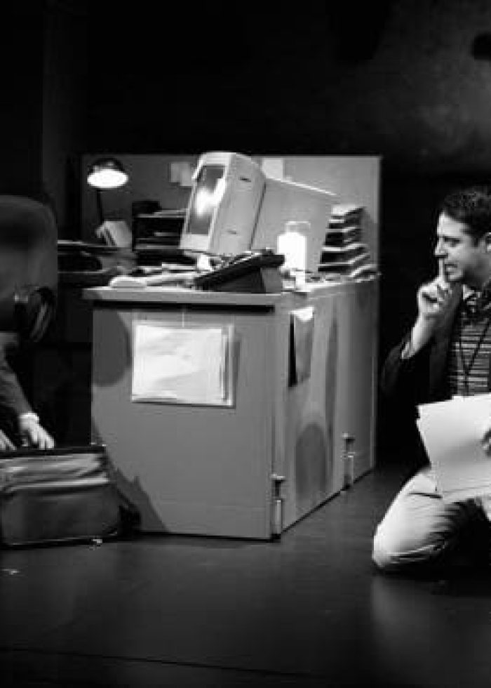 Actors in "CQ/CX" kneeling on stage near a desk, one on the phone, the other with a briefcase.