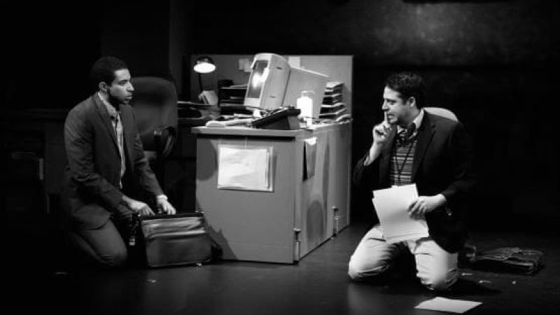 Actors in "CQ/CX" kneeling on stage near a desk, one on the phone, the other with a briefcase.