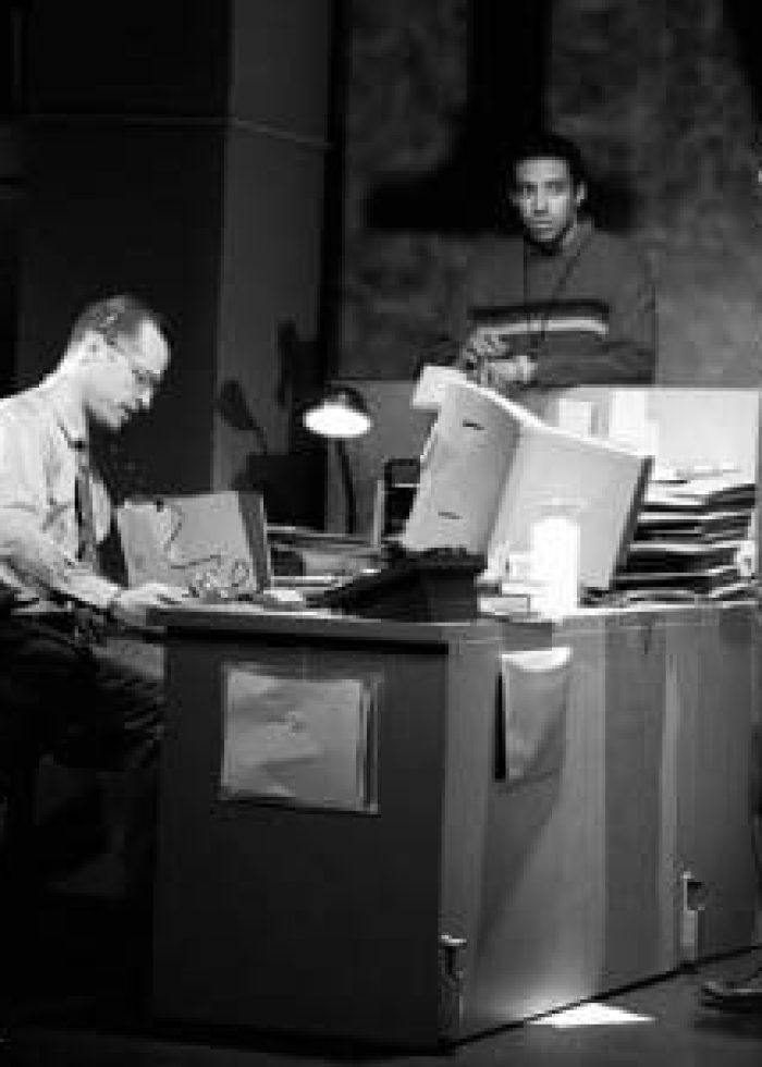 Three men in an office, one seated, two standing, discussing work. "CQ/CX" review.