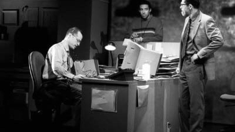 Three men in an office, one seated, two standing, discussing work. "CQ/CX" review.