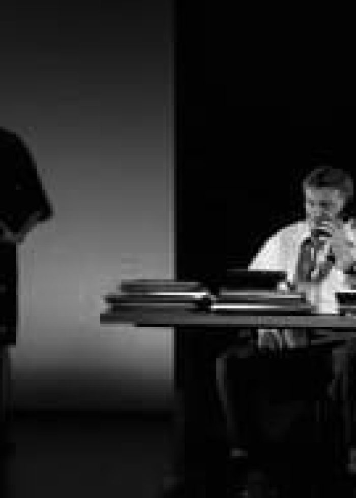 Two actors in a play; one on the phone, the other at a desk with files.