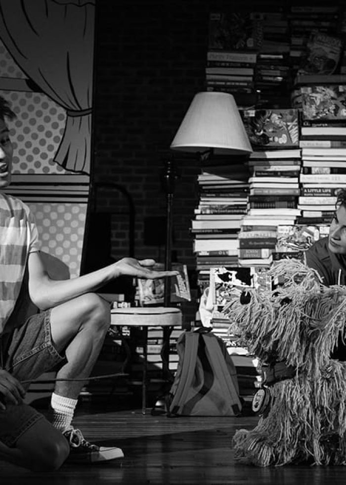 Two actors on stage with a puppet dog, part of a theatrical production.