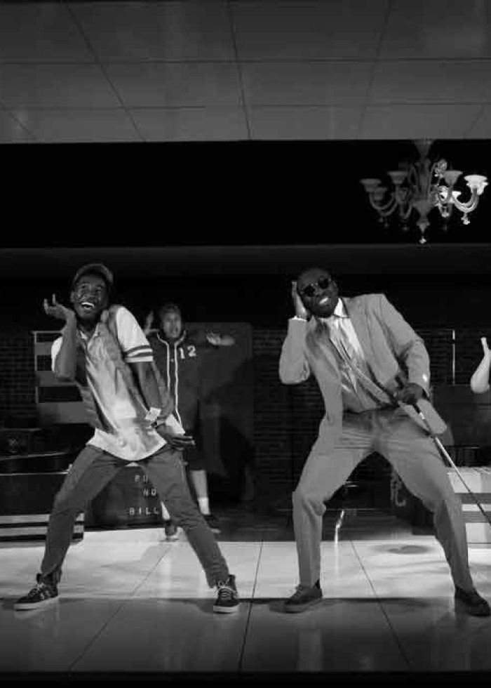 Actors performing a dance on stage. A man in a suit leads the group. Black and white photo.