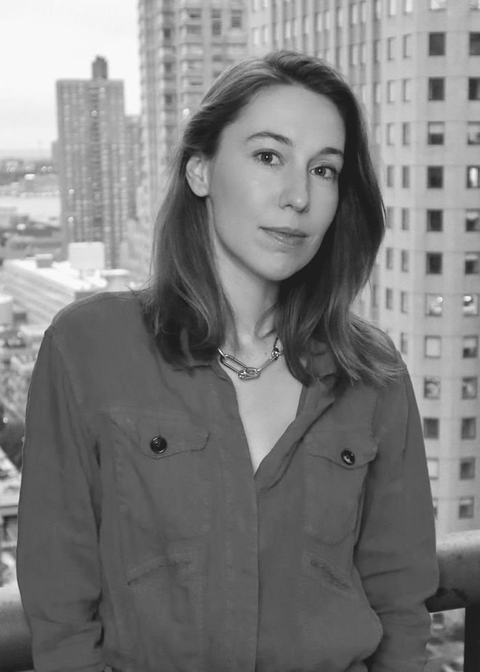 Mona Pirnot posing in front of a cityscape wearing a red shirt and silver chain necklace.