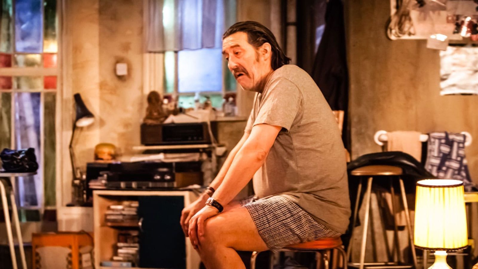 Man in undershirt and shorts sits on stool, looking pensive. The Night Alive.