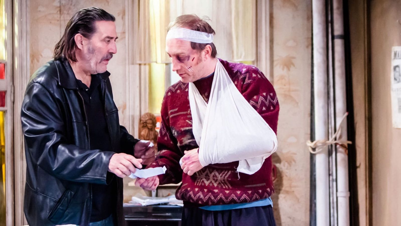 Two men, one injured, exchange a note in a dimly lit room. The Night Alive.