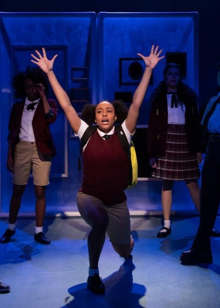Cast of "She Persisted" musical on stage. A woman kneels center, arms raised, surrounded by other performers.