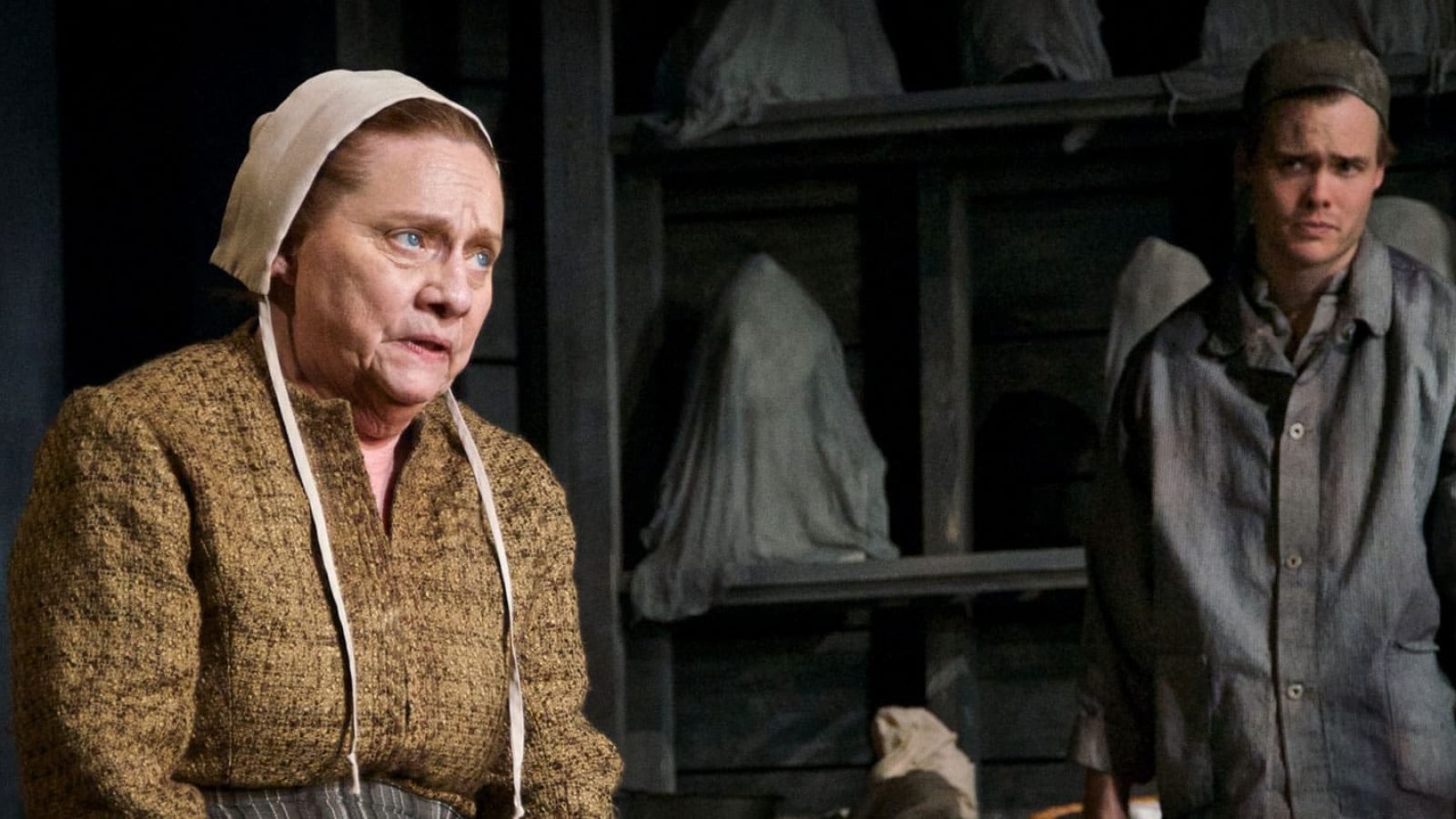 Two figures in period costume, a woman and a man, stand before a shelf of shrouded objects. A skeleton is visible in the background.