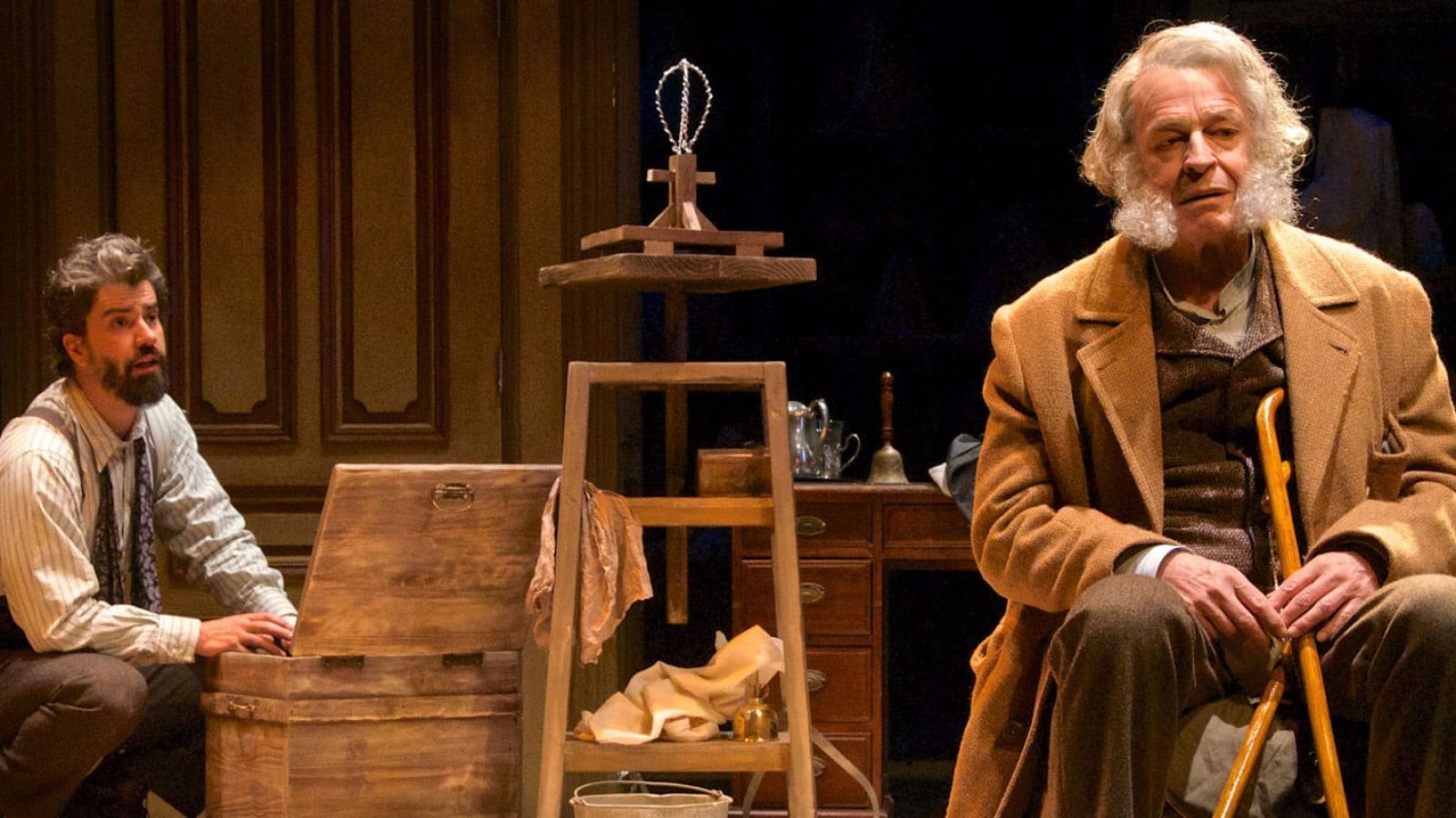 Two men in period costume; one seated, holding a cane, the other kneeling. A wooden chest and antique furniture are in the background.