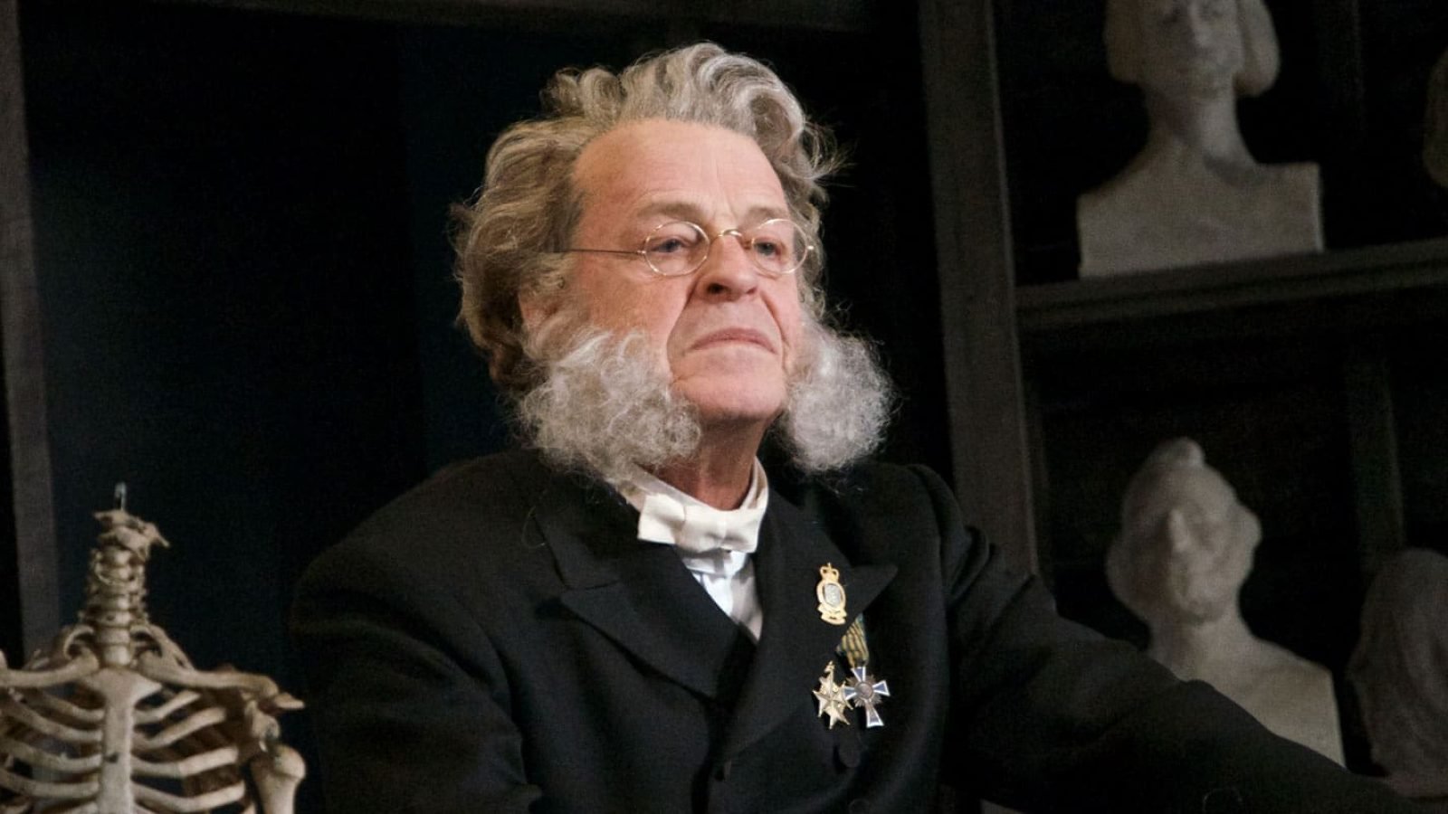 Elderly man with long white hair and beard, wearing a dark suit and medals, sits amidst plaster busts.