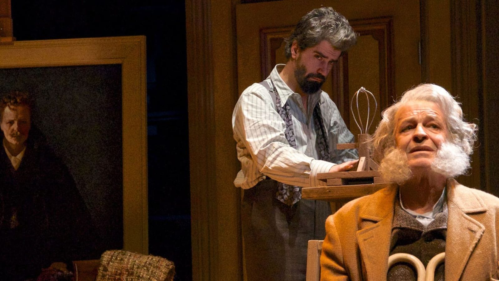 Two men in period clothing, one older with a long beard, the other younger, in an artist's studio. A painting hangs on an easel in the background.