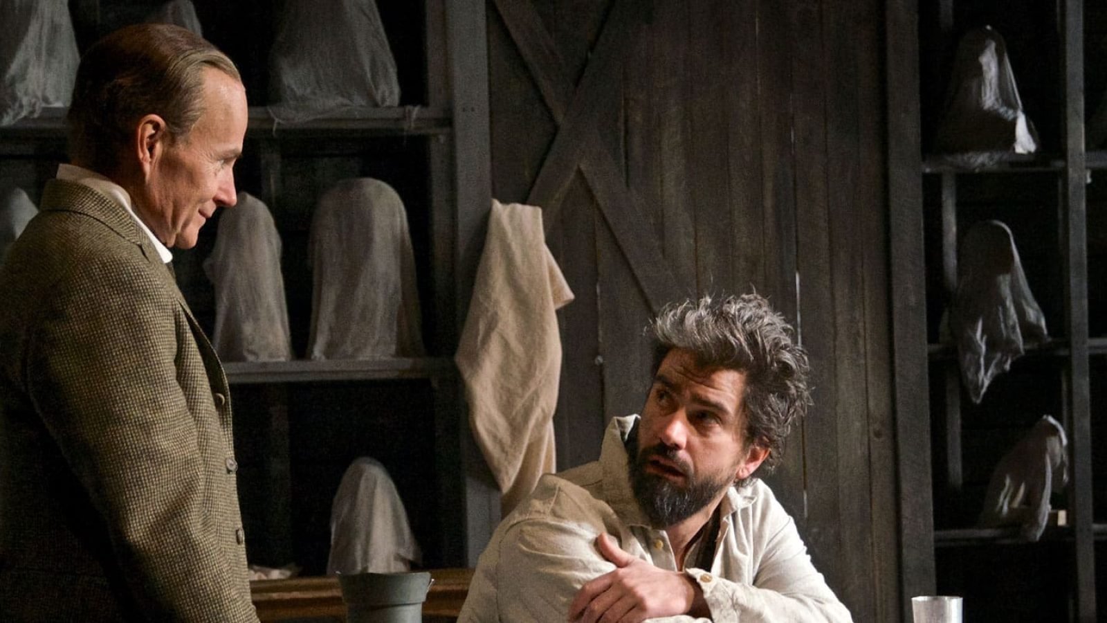 Two men converse in a dimly lit room filled with shrouded busts and a skeleton.
