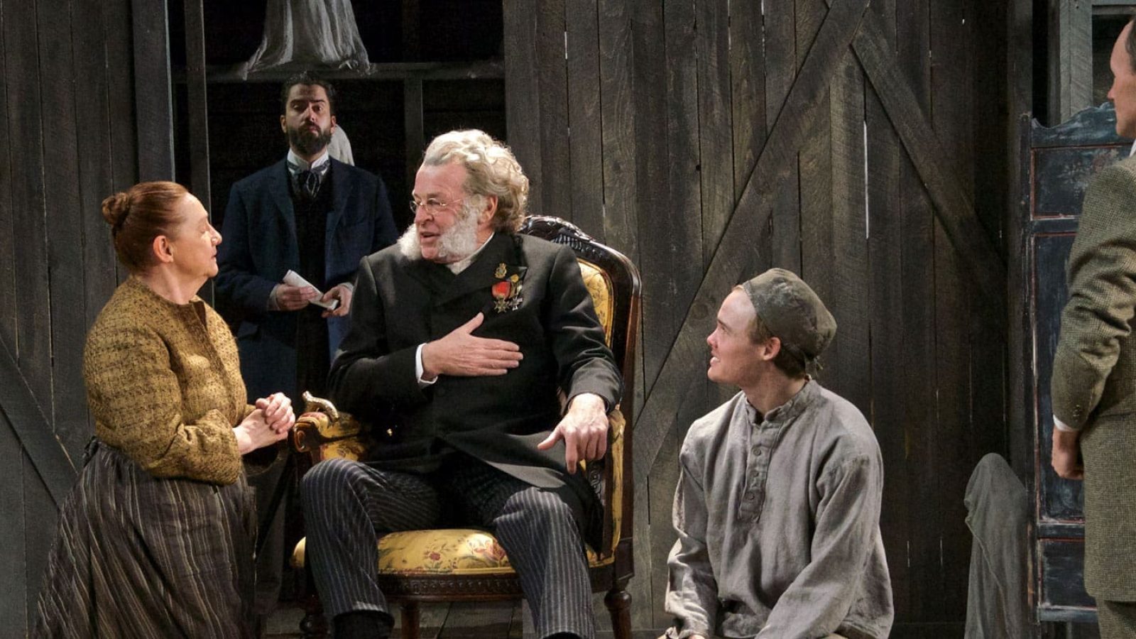 A family scene, possibly from a play, depicting a seated older man in period clothing, surrounded by family members. The image evokes themes of legacy and posterity.