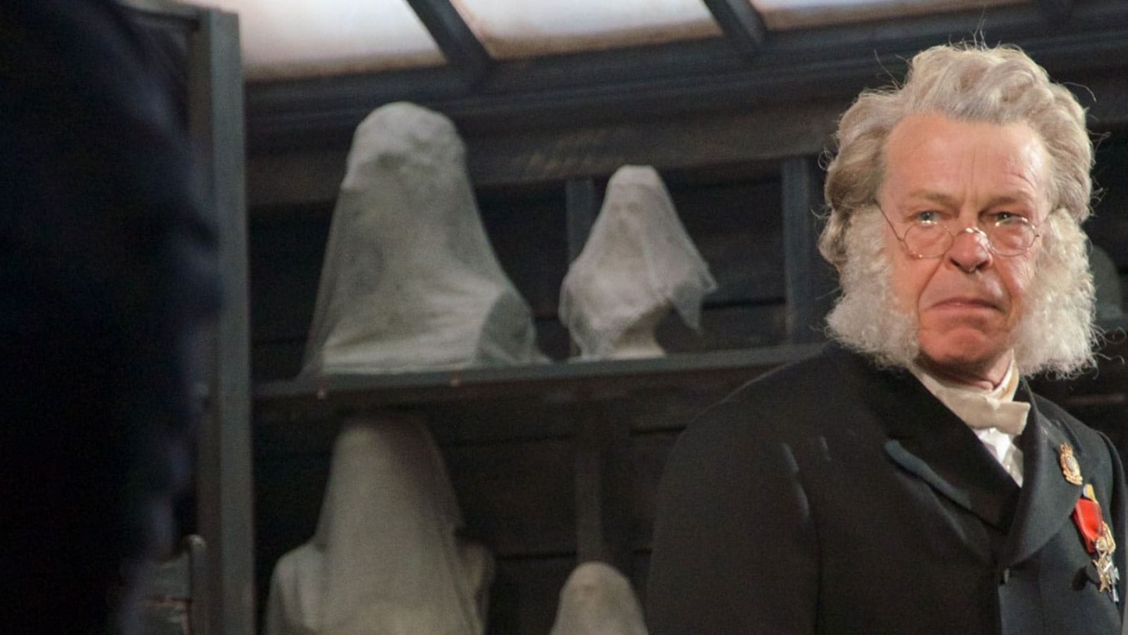 Older man with white hair and beard, wearing glasses and a dark coat, stands in front of covered busts. Posterity.