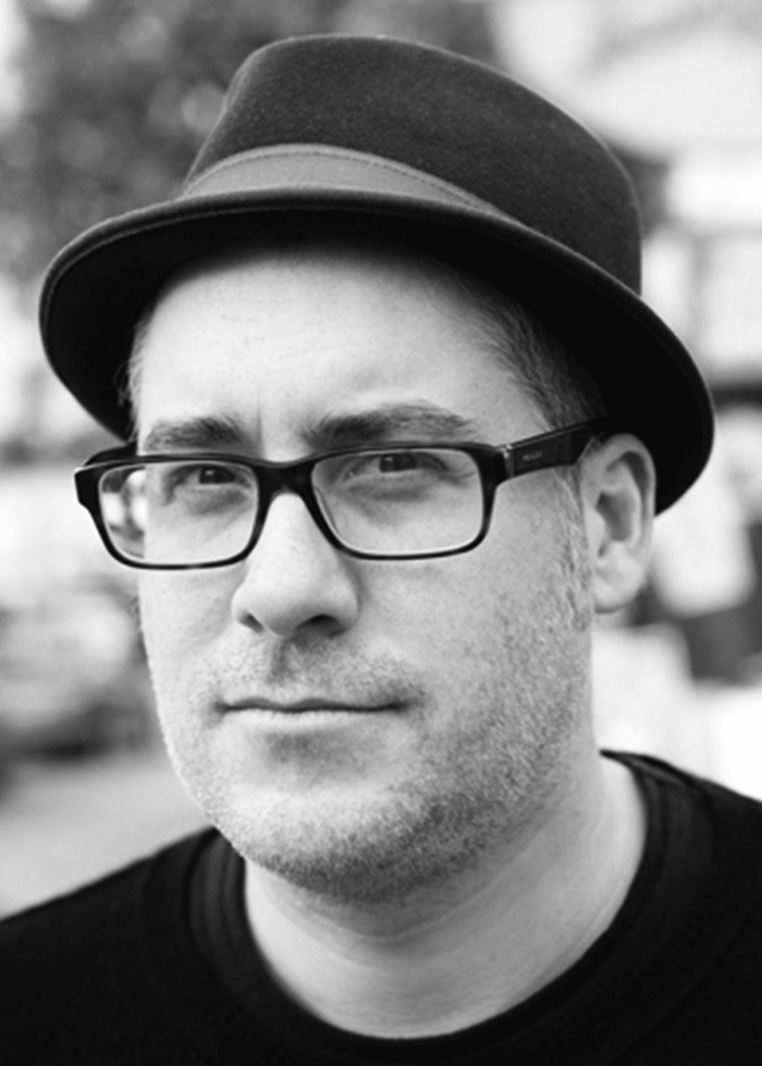 Rolin Jones wearing a fedora and glasses, looking at the camera